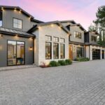 Modern luxury home exterior with large windows, stone facade, and spacious driveway at sunset.